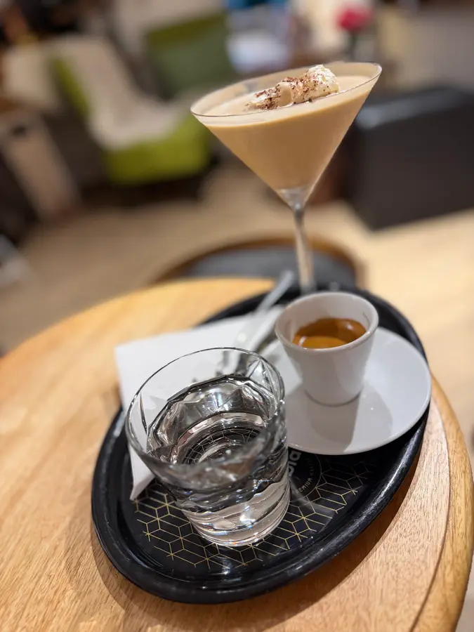 Espresso mit Wasserglas und ein cremiges Getränk im Cocktailglas auf einem runden Holztisch