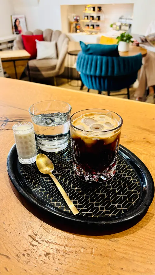 Eiskaffee im Glas mit goldenem Löffel, dahinter ein Samtsessel in Petrol
